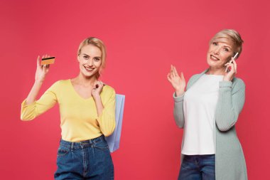 happy woman holding credit card and shopping bag near mother talking on smartphone isolated on pink