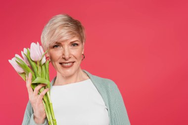 stylish woman smiling at camera while holding fresh tulips isolated on pink