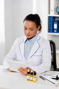 Beyaz önlüklü Afro-Amerikan doktor CBI 'lı şişelerin yanında dijital tablet kullanıyor.