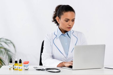 Afro-Amerikalı doktor masadaki tıbbi esrar ve CBD şişelerinin yanında dizüstü bilgisayar kullanıyor.