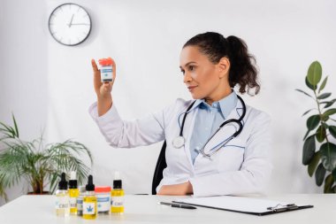 Steteskoplu esmer Afrikalı Amerikalı doktor klinikte üzerinde tıbbi esrar yazan şişeye bakıyor.