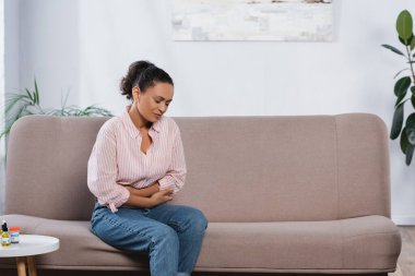 african american woman suffering from abdominal pain in living room  clipart