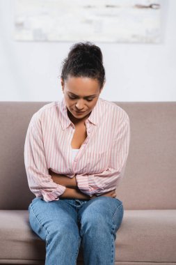 Afro-Amerikan kadın oturma odasında kanepede otururken karın ağrısı çekiyor. 