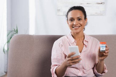 Gülümseyen Afrikalı Amerikalı kadın elinde tıbbi esrar ve akıllı telefon şişesi tutuyor.