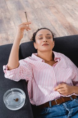 high angle view of african american woman lying on sofa and smoking rolled joint with medical cannabis near ashtray  clipart