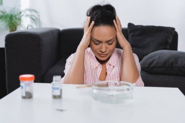 Afrika kökenli Amerikalı kadın, bulanık ön planda tıbbi esrar bulunan şişelerin yanında migren ağrısı çekiyor.