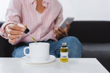 Afro-Amerikan kadının elinde akıllı telefon ve cbd sıvısıyla çay bardağının yanında pipetle görüntüsü.