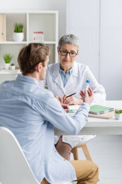 Doktor defterlerin yanında oturuyor ve hasta ön planda aşıyı tutuyor. 