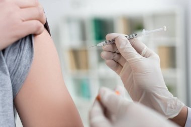 Cropped view of doctor in latex gloves holding syringe and vaccine on blurred foreground near patient  clipart