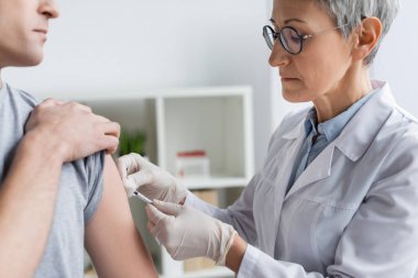 Gözlüklü olgun bir doktor hastanede ön planda bulanık bir adama aşı yapıyor. 