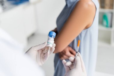 Doktorun elinde koronavirüs aşısı ve şırıngayla arka planda bulanık bir kadın görüntüsü. 
