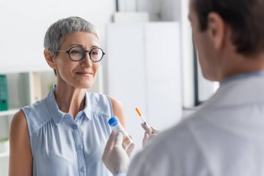 Gülümseyen kadın önplanda şırınga ve aşıyla doktora bakıyor. 
