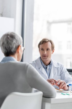 Beyaz önlüklü doktor aşının yanında hastayla konuşuyor ve ön planda bulanık haplar var. 