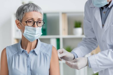 Tıbbi maskeli olgun bir kadın ve klinikte aşı yapan bir doktor. 