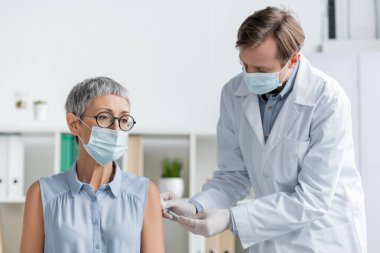 Lateks eldivenli ve tıbbi maskeli bir doktor hastanede yetişkin bir hastaya aşı yapıyor. 