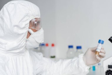 Scientist in protective suit and goggles holding vaccine on blurred background  clipart