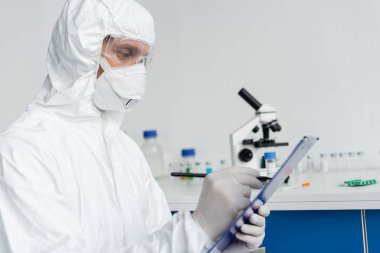 Scientist in protective uniform writing on clipboard on blurred foreground  clipart