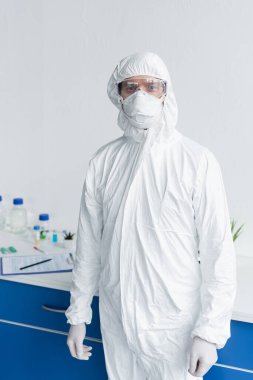 Tehlikeli madde giysisi ve gözlüklü bilim adamları laboratuvarda kameraya bakıyor. 