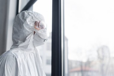 Tıbbi maskeli, gözlüklü ve koruyucu giysili bilim adamı pencerenin yanında duruyor. 