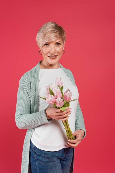 зрелая, стильная женщина, держащая свежие тюльпаны, глядя на камеру, изолированную на розовый