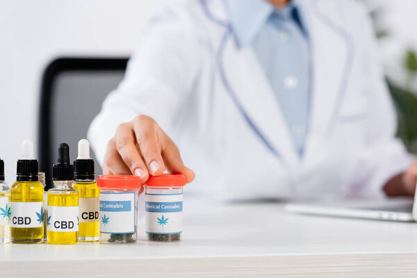 cropped view of blurred doctor reaching bottles with medical cannabis and cbd lettering on table 