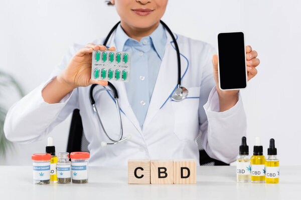cropped view of african american doctor holding blister pack with capsules and smartphone with blank screen near cbd cubes and bottles 