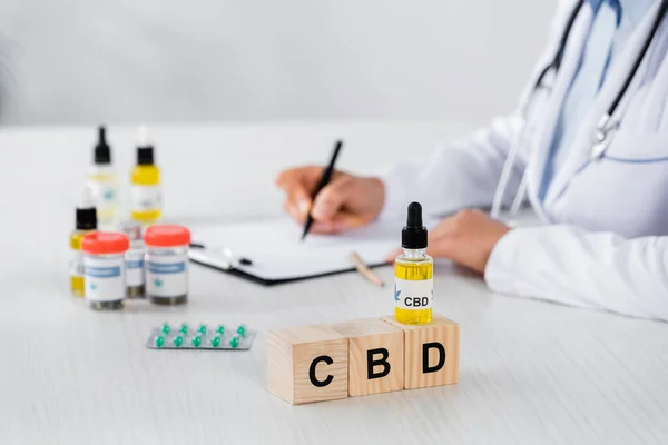 partial view of doctor writing prescription near cbd cubes and medication on desk