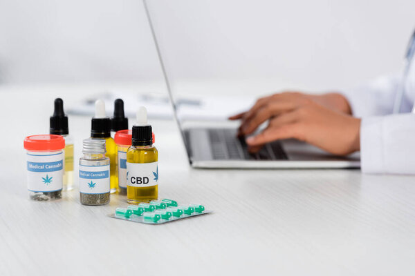 cropped view of african american doctor typing on laptop near cannabis medication on desk