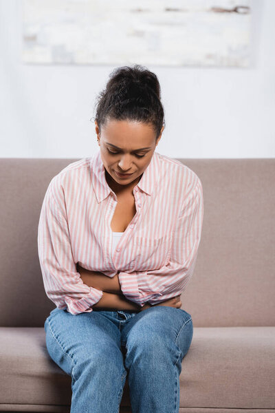 african american woman suffering from abdominal pain while sitting on sofa in living room 