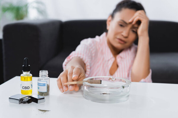 blurred african american woman suffering from migraine and reaching joint in astray near bottles with medical cannabis and cbd on table