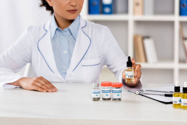 cropped view of african american doctor in white coat holding bottle with cbd lettering in clinic
