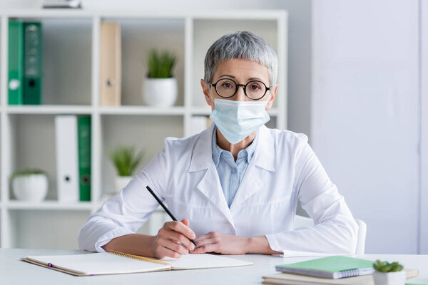 Mature doctor in medical mask holding pen near notebooks on blurred foreground 