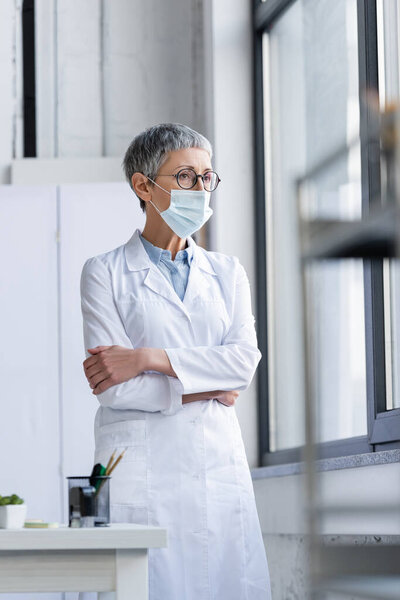 Mature doctor in white coat and medical mask standing near window and table on blurred foreground 