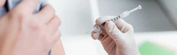 Cropped view of doctor in latex glove holding syringe near patient on blurred foreground, banner 