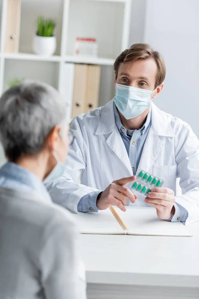 Doctor holding pills near notebook and patient in medical mask on blurred foreground