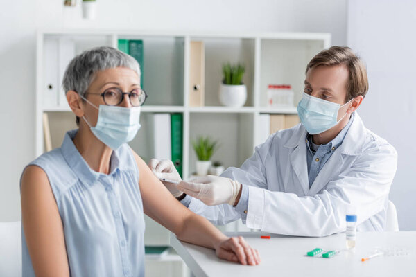 Doctor in medical mask doing vaccination of mature patient on blurred foreground near pills on table 