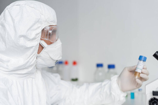 Scientist in protective suit and goggles holding vaccine on blurred background 