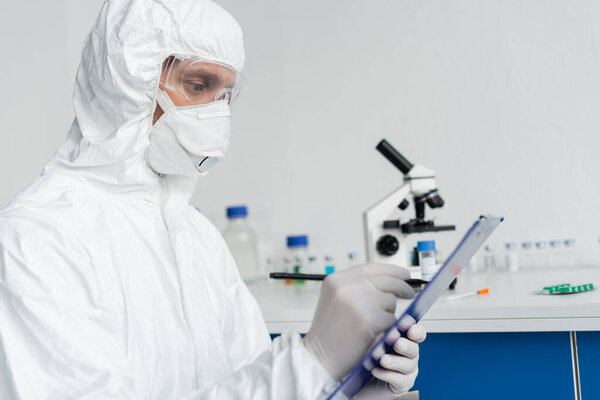 Scientist in protective uniform writing on clipboard on blurred foreground 