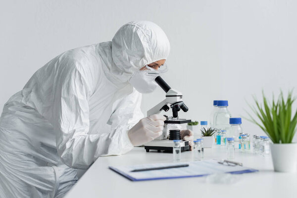 Scientist in protective uniform using microscope near vaccines and clipboard on blurred foreground 