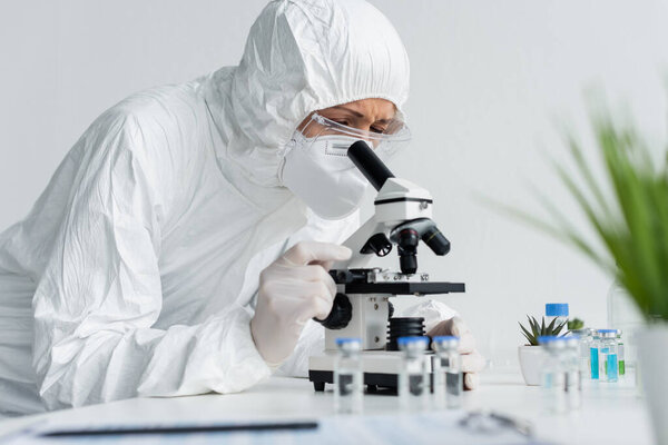 Scientist in hazmat suit working with microscope near vaccines on blurred foreground 