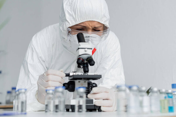 Scientist in goggles and hazmat suit looking at microscope near vaccines on blurred foreground 