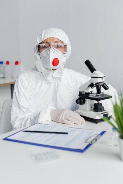 Scientist looking at camera near microscope and clipboard on blurred foreground 