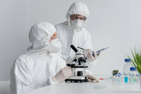 Scientist using microscope near colleague with clipboard and vaccines 