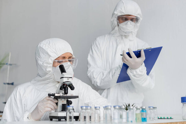 Scientist in protective uniform using microscope near colleague and vaccines on blurred foreground 