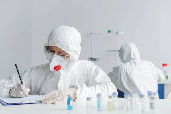 Scientist Protective Uniform Writing Clipboard Vaccines Colleague Blurred Background — Φωτογραφία Αρχείου