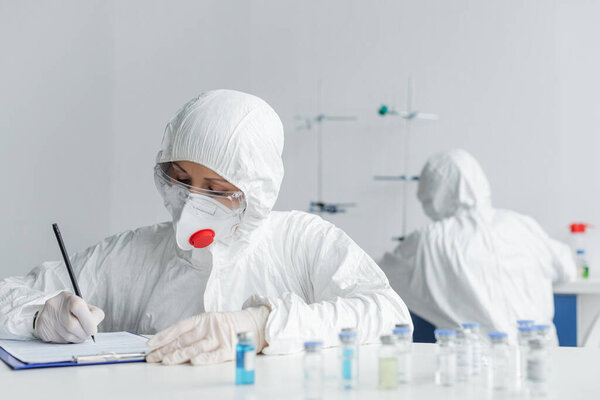 Scientist in protective uniform writing on clipboard near vaccines and colleague on blurred background 