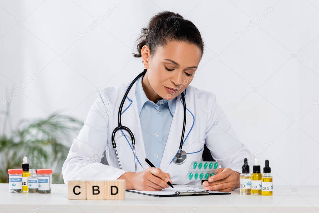 african american doctor with stethoscope holding blister pack with capsules near cbd cubes and bottles while writing prescription 