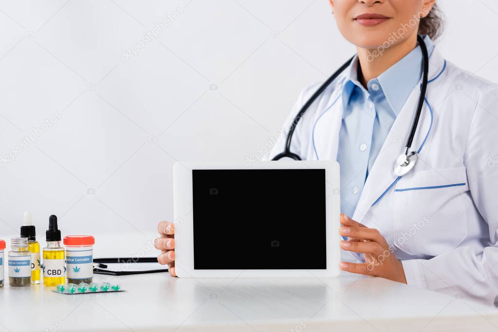 partial view of african american doctor holding digital tablet with blank screen near cannabis medication on desk