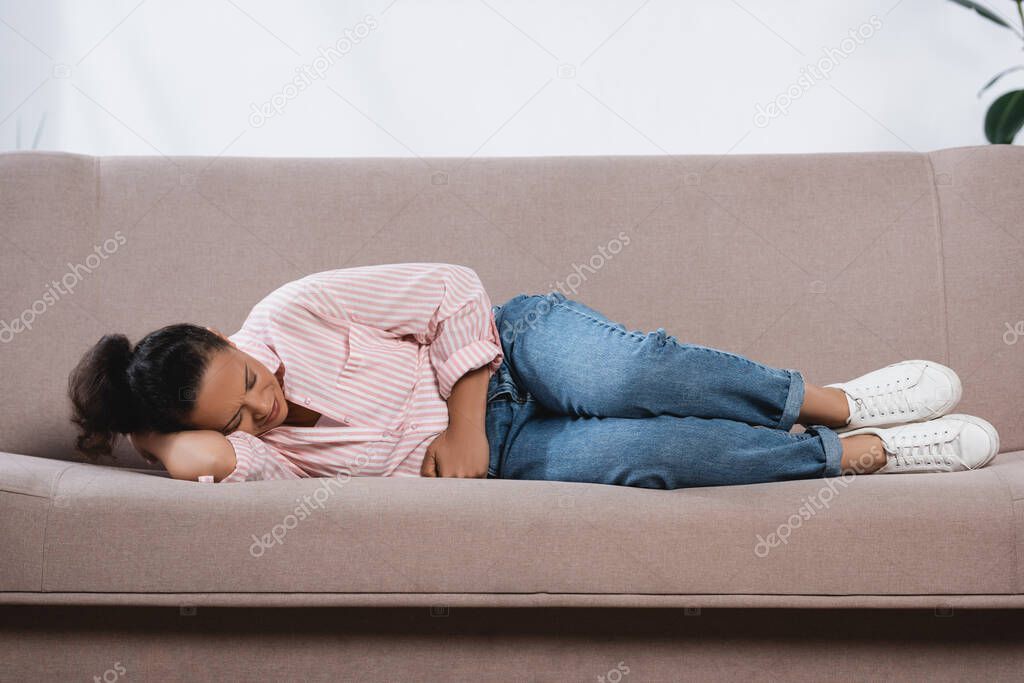 african american woman suffering from stomach ache while lying on sofa in living room 