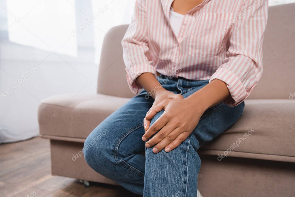 cropped view of african american woman suffering from knee pain while sitting on couch